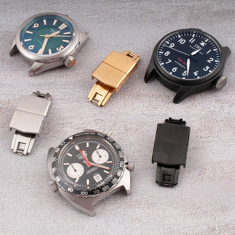 Spring-loaded deployant buckles for CTS straps in silver, gold, and black PVD finishes displayed with watches on a gray surface